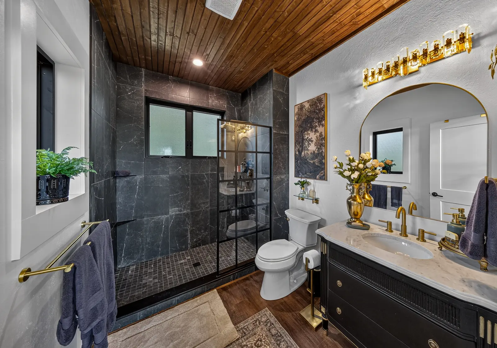The bath with a black-framed glass walk-in shower, arched mirror and brass vanity lighting